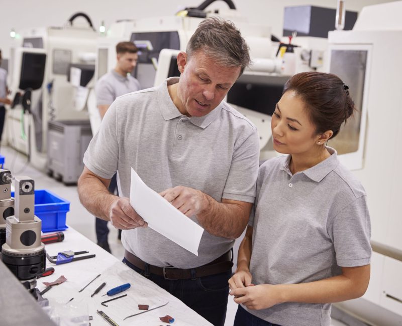 Engineer And Female Apprentice Discussing Job Sheet In Factory